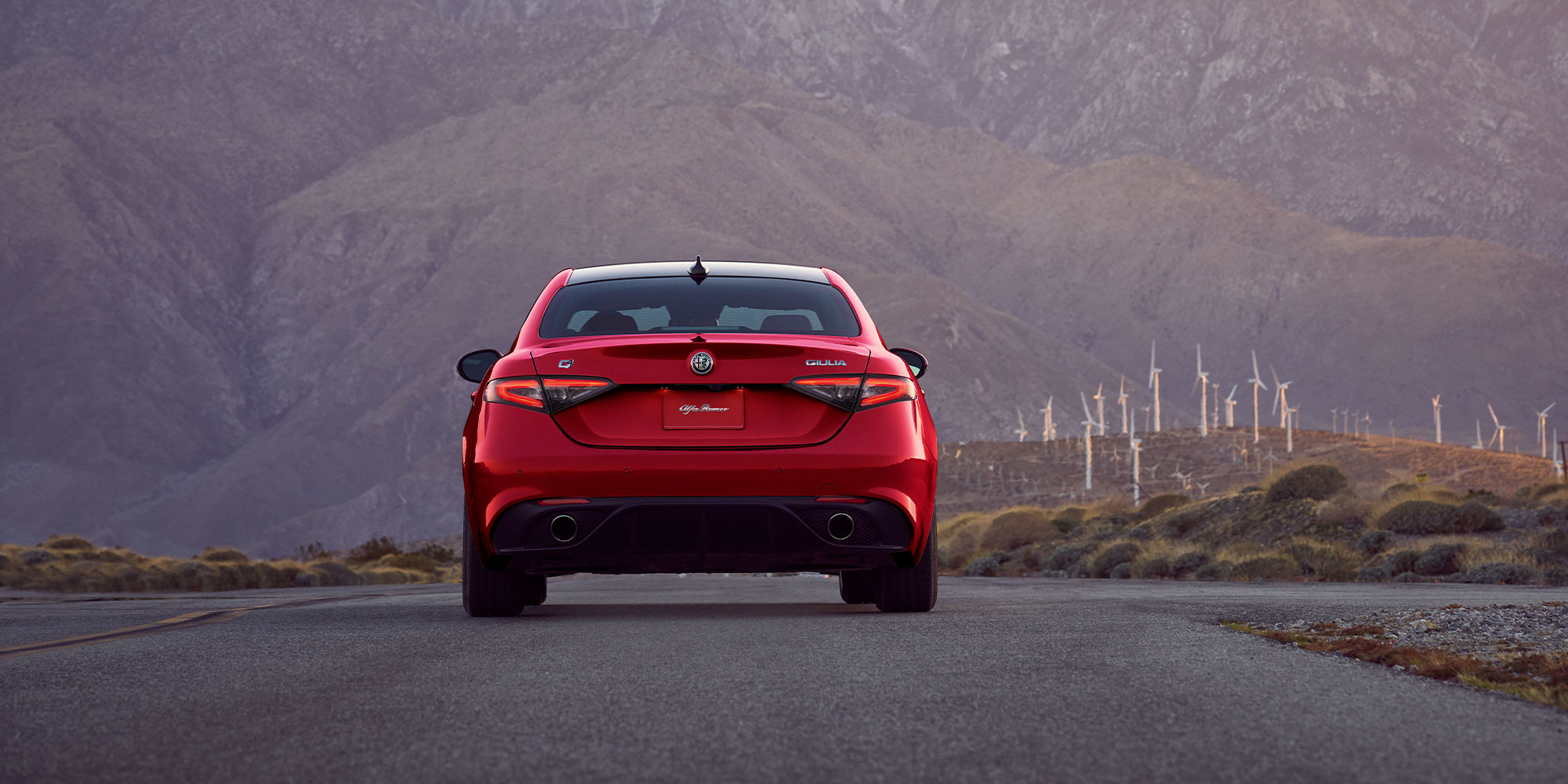 Rear view of an Alfa Rosso 2025 Alfa Romeo Giulia on a road with wind turbines and mountains in the distance.