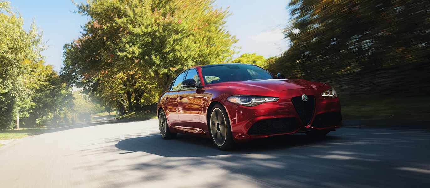 2025 Alfa Rosso Alfa Romeo Giulia, driving under a clear sky, on a road surrounded by lush green trees.