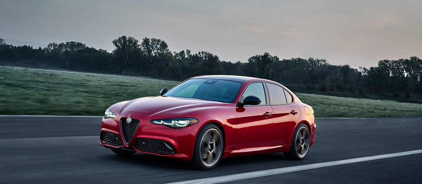 2025 Alfa Rosso Alfa Romeo Giulia, driving along a road in the early morning, with a green field and trees in the distance.