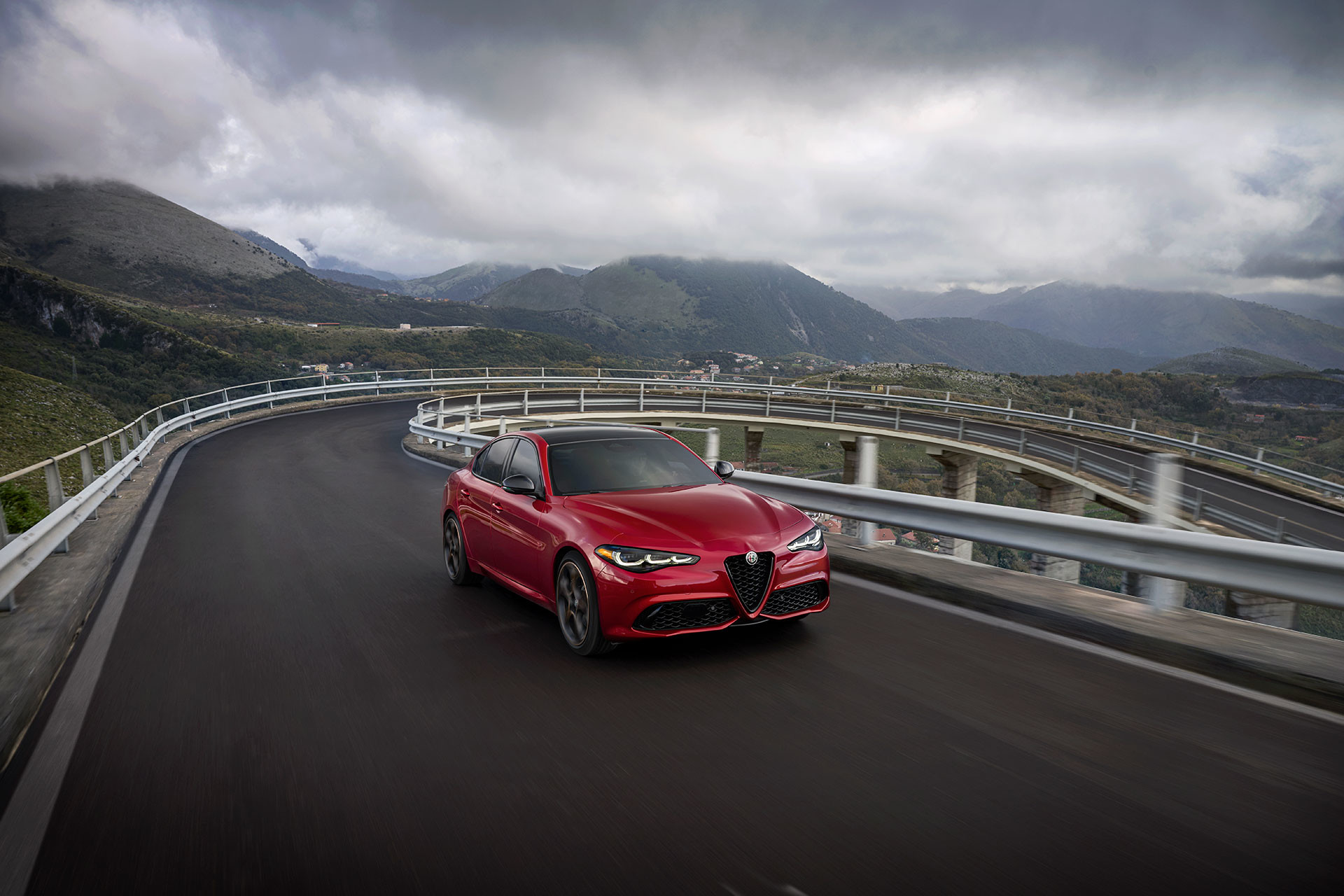 Red 2026 Alfa Romeo Giulia driving on a scenic mountain road with cloudy skies.