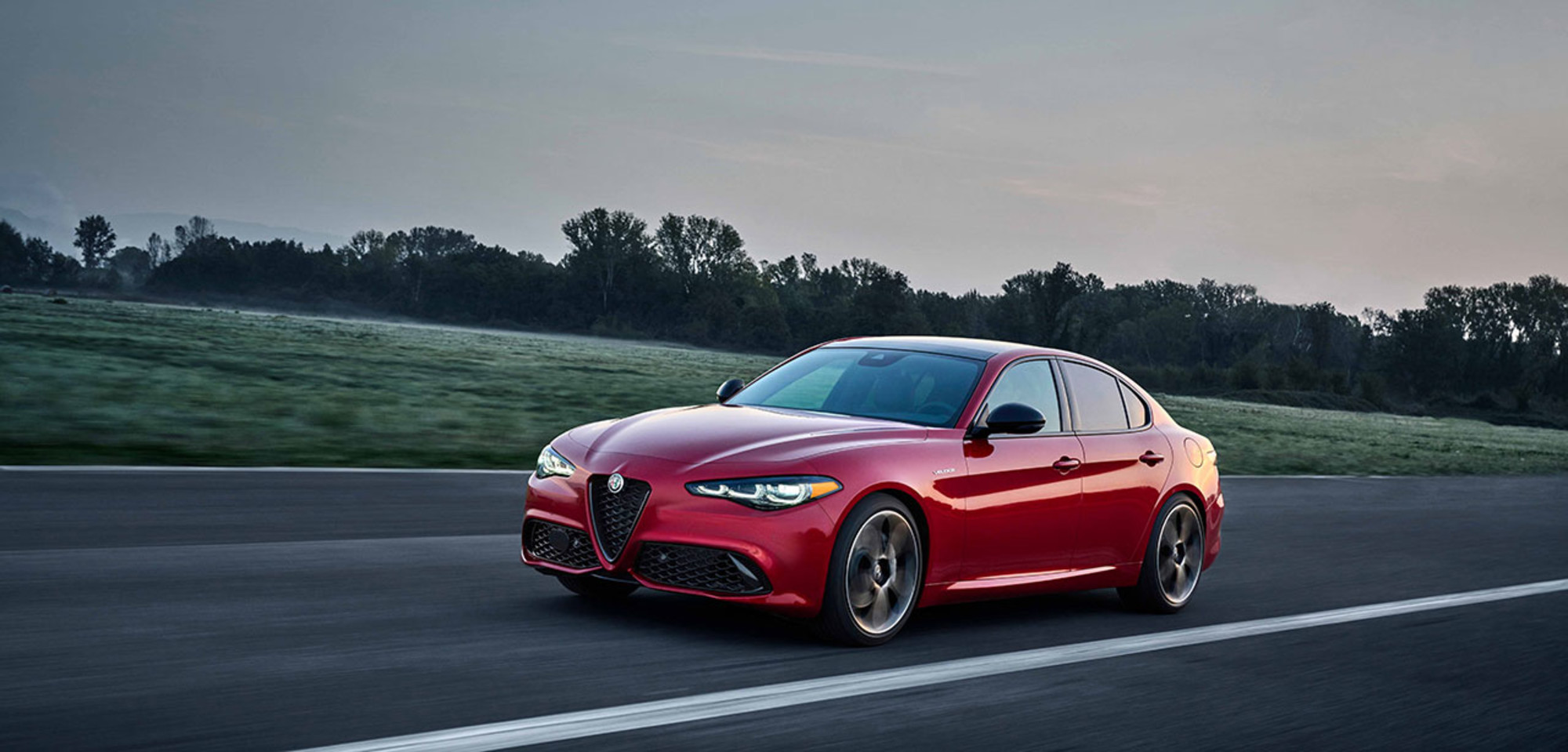 Red 2026 Alfa Romeo Giulia driving on a straight road with a scenic backdrop.