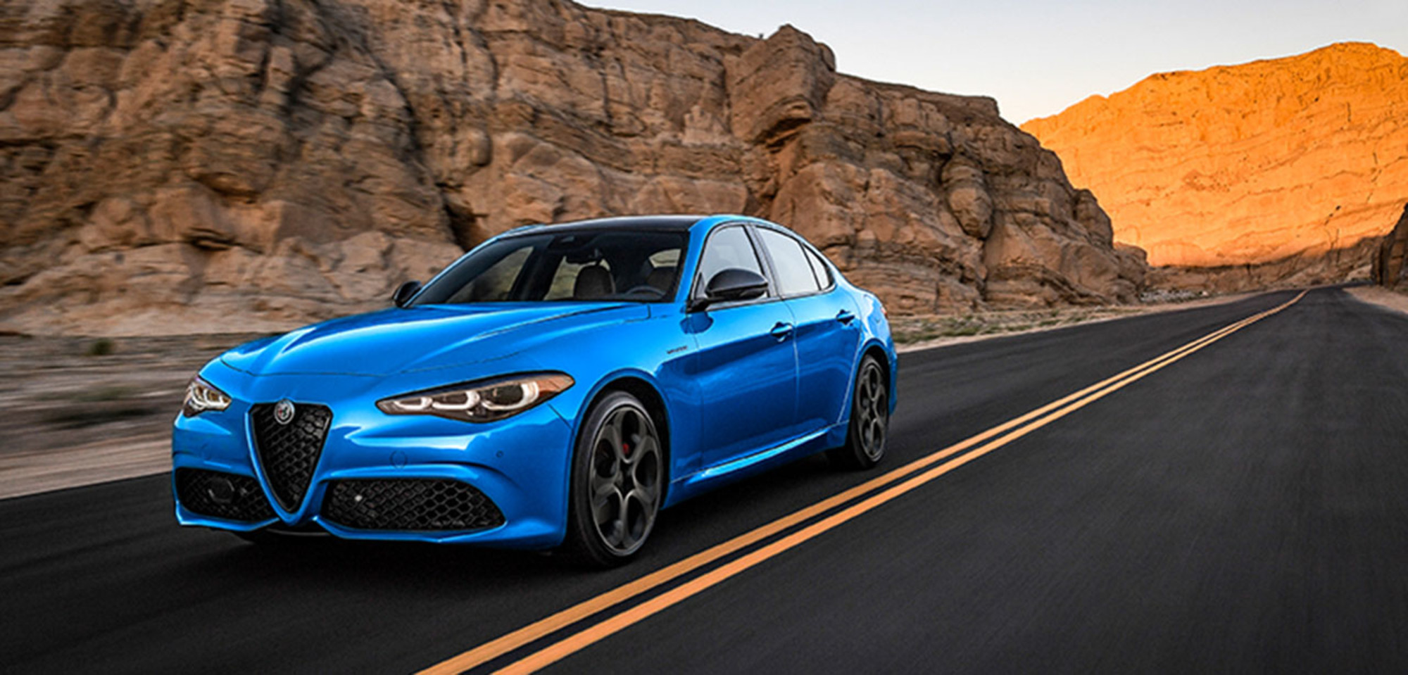 Blue 2026 Alfa Romeo Giulia driving through a desert canyon landscape at sunset.
