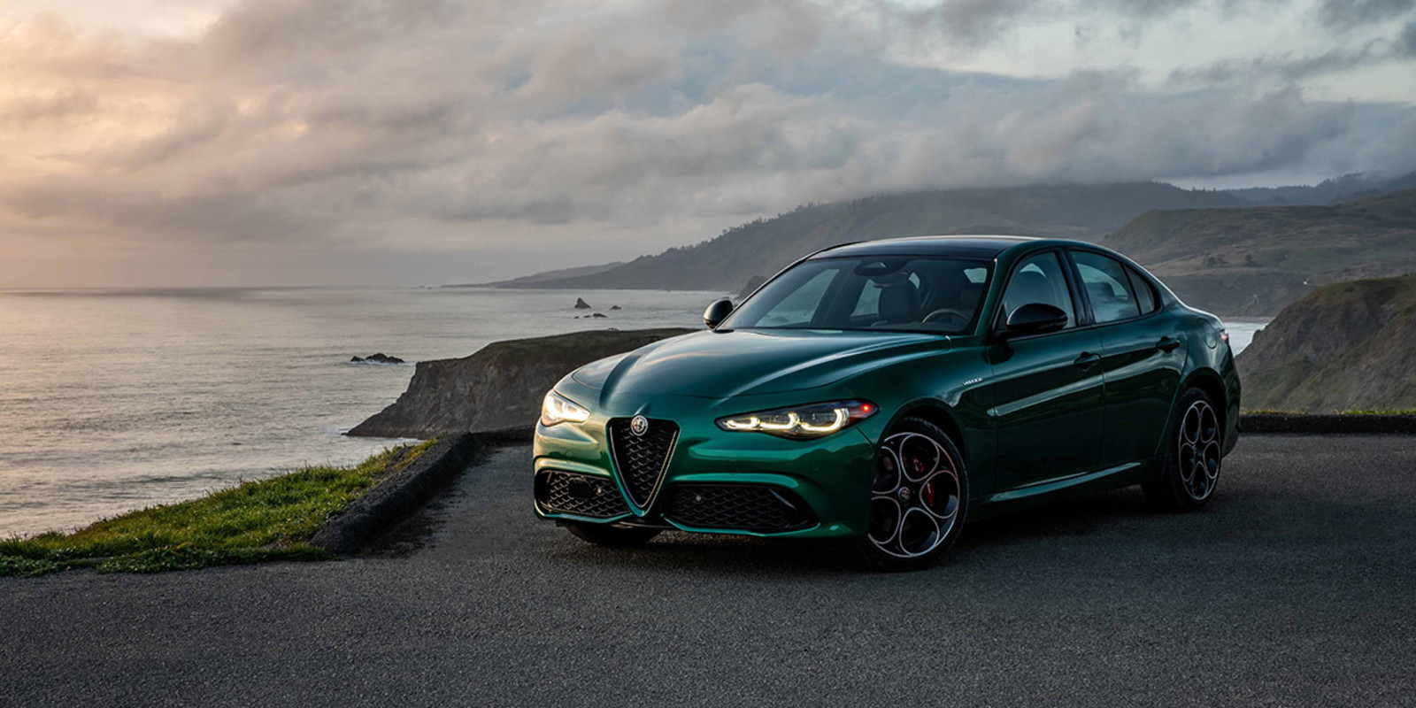 A green 2026 Alfa Romeo Giulia parked on a cliffside road overlooking the ocean at sunset.
