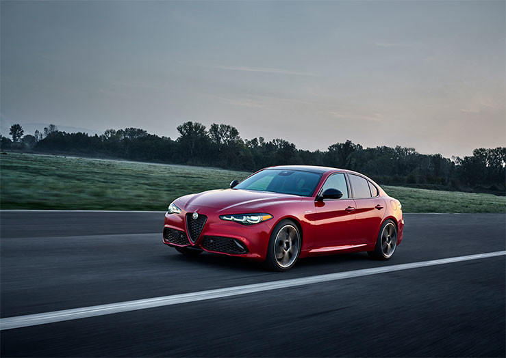 Une Alfa Romeo Giulia rouge 2026 roule sur une route droite avec un décor pittoresque en arrière-plan.