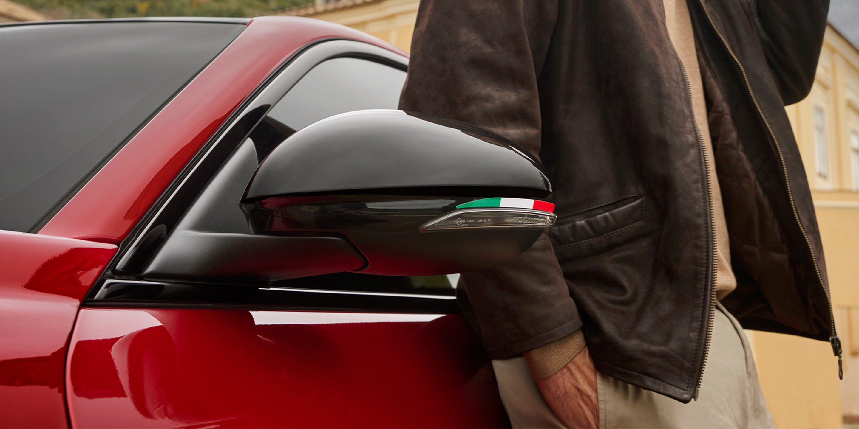 Un gros plan du rétroviseur latéral d’une voiture Alfa Romeo rouge avec un fini noir élégant et un motif de drapeau italien. 