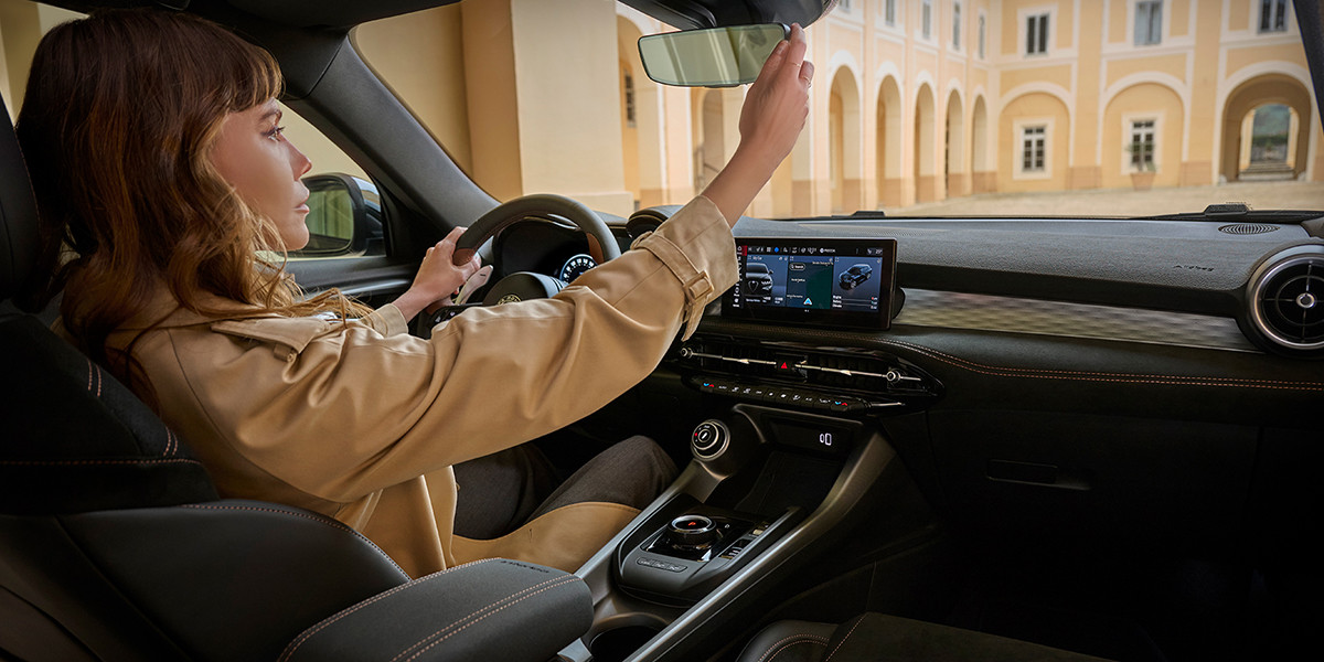 Une personne assise à l'intérieur d'une Alfa Romeo Intensa ajuste le rétroviseur. L'intérieur est doté d'un tableau de bord moderne avec écran tactile.