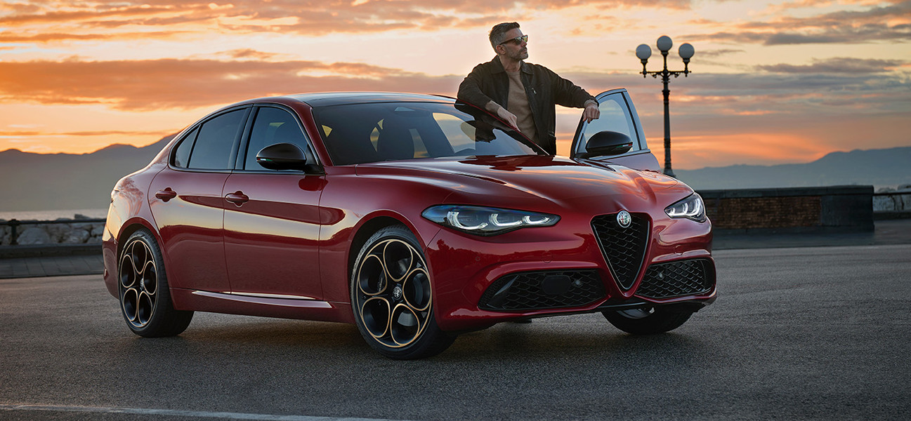Une Alfa Romeo Giulia rouge est stationnée au bord de la route pendant le coucher du soleil. Une personne se tient debout près de la porte latérale du côté conducteur ouverte en regardant vers l’horizon.