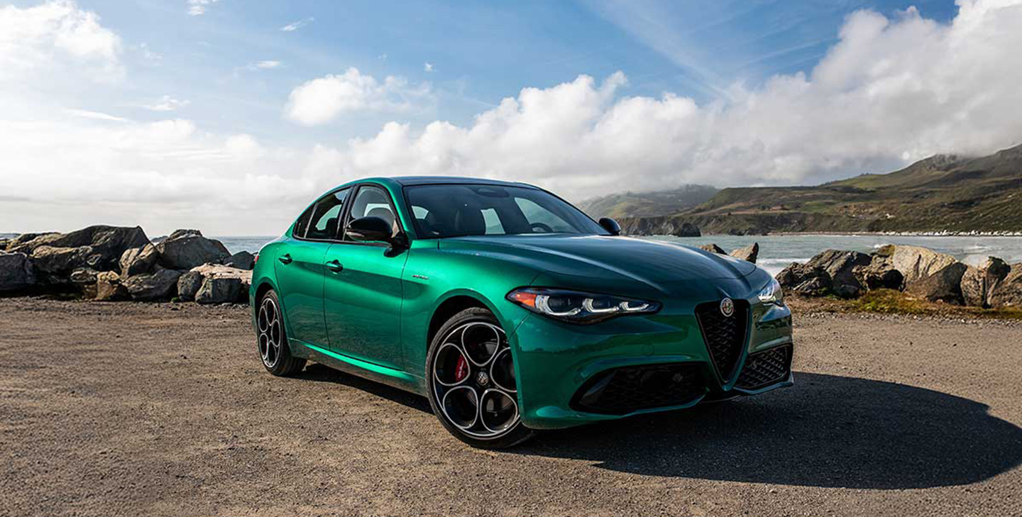 Front three-quarter view of a metallic green Alfa Romeo Giulia sedan parked on a gravel coastal overlook with the ocean in the background.
