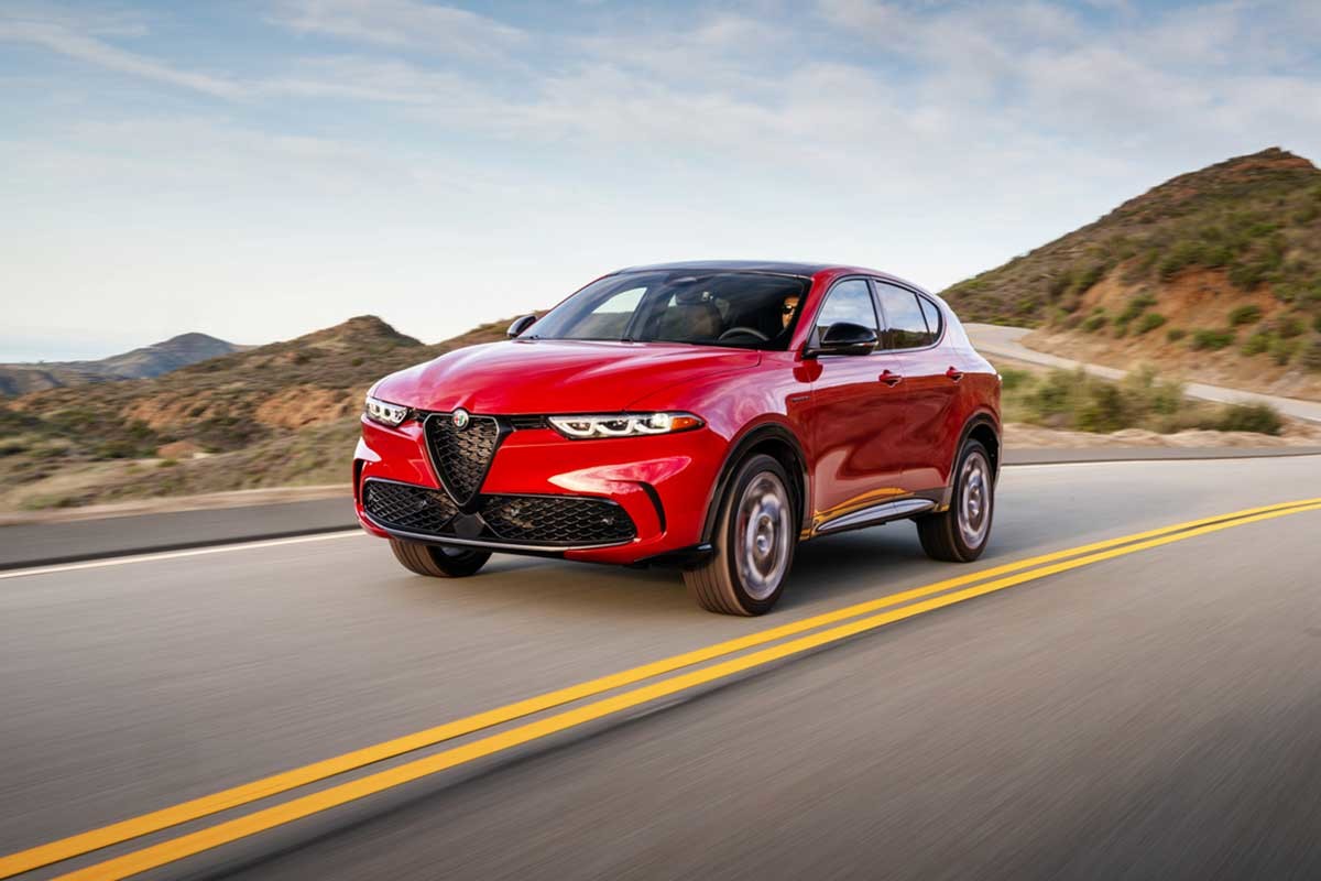 A red 2026 Alfa Romeo Stelvio driving on a road with a blue sky and mountains in the background.