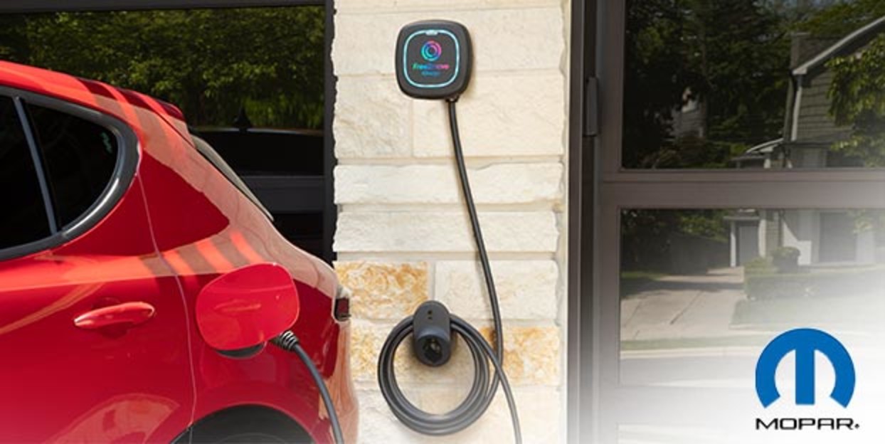 Red 2025 Alfa Romeo electric vehicle plugged into a wall-mounted EV charger on the exterior of a modern home. Buy Now From Mopar<small>®</small>