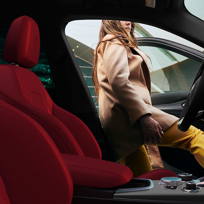 Un profil latéral de la vue du passager montrant une femme prenant place dans le siège conducteur d'un Stelvio 2022 avec des sièges en cuir rouge et une finition noire