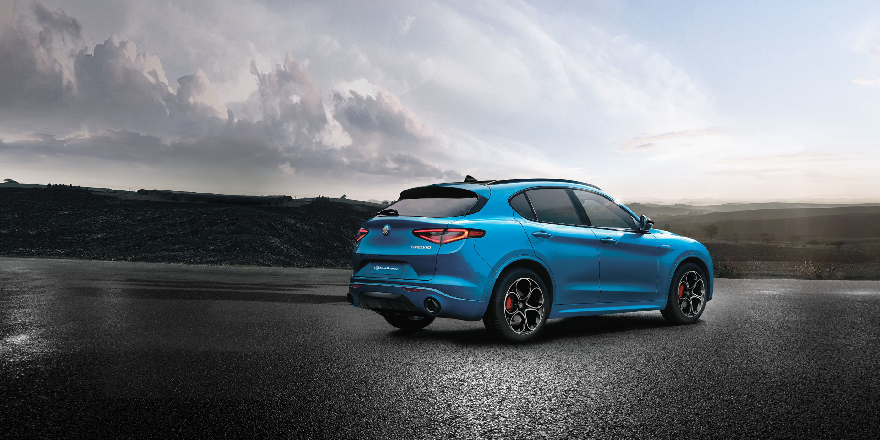 Front close-up view of blue 2023 Alfa Romeo Stelvio driving on the road with rock mountains in the background.



