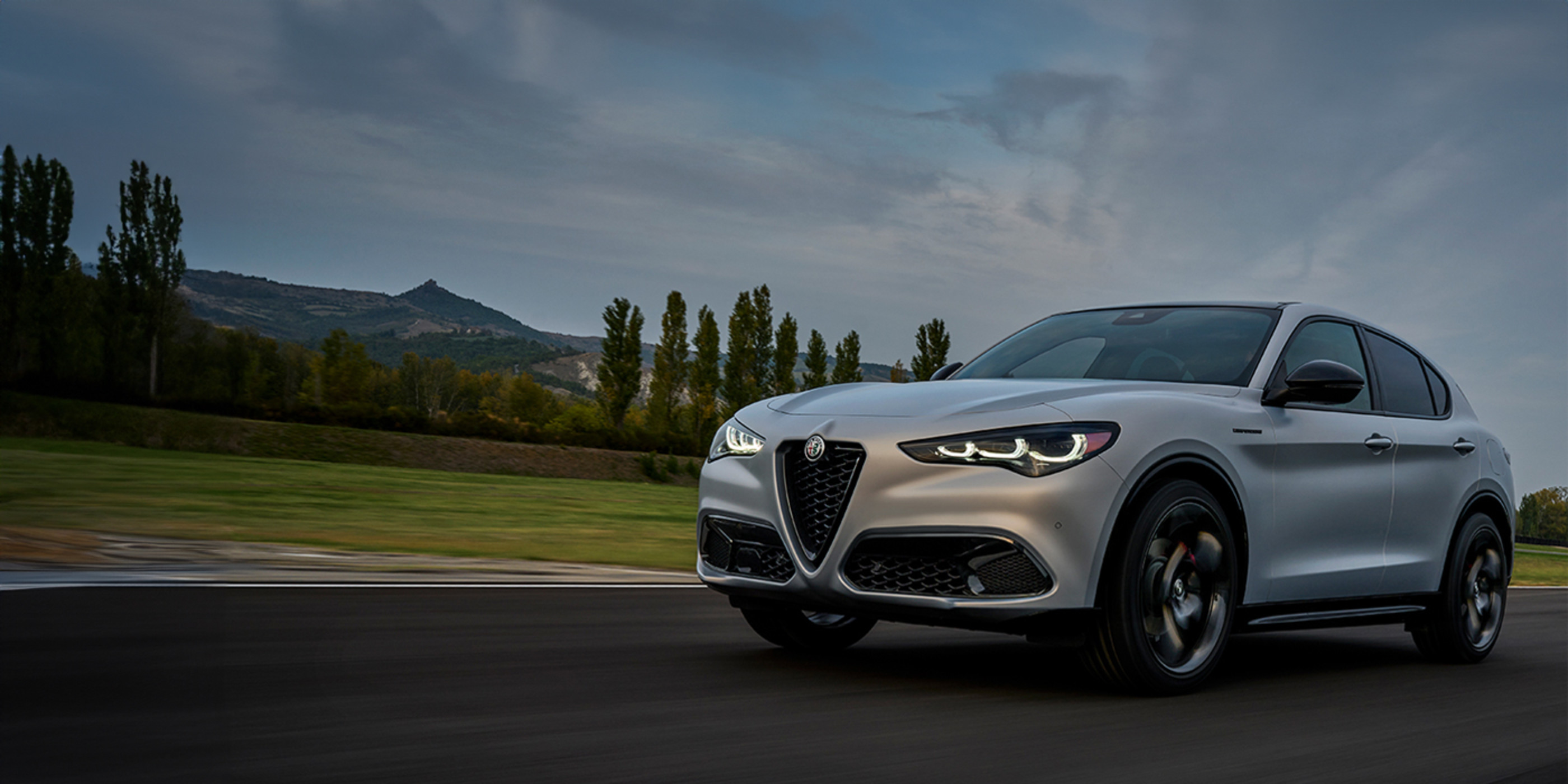 A front angled view of a silver 2024 Alfa Romeo Stelvio driving on a paved road with trees shown behind.