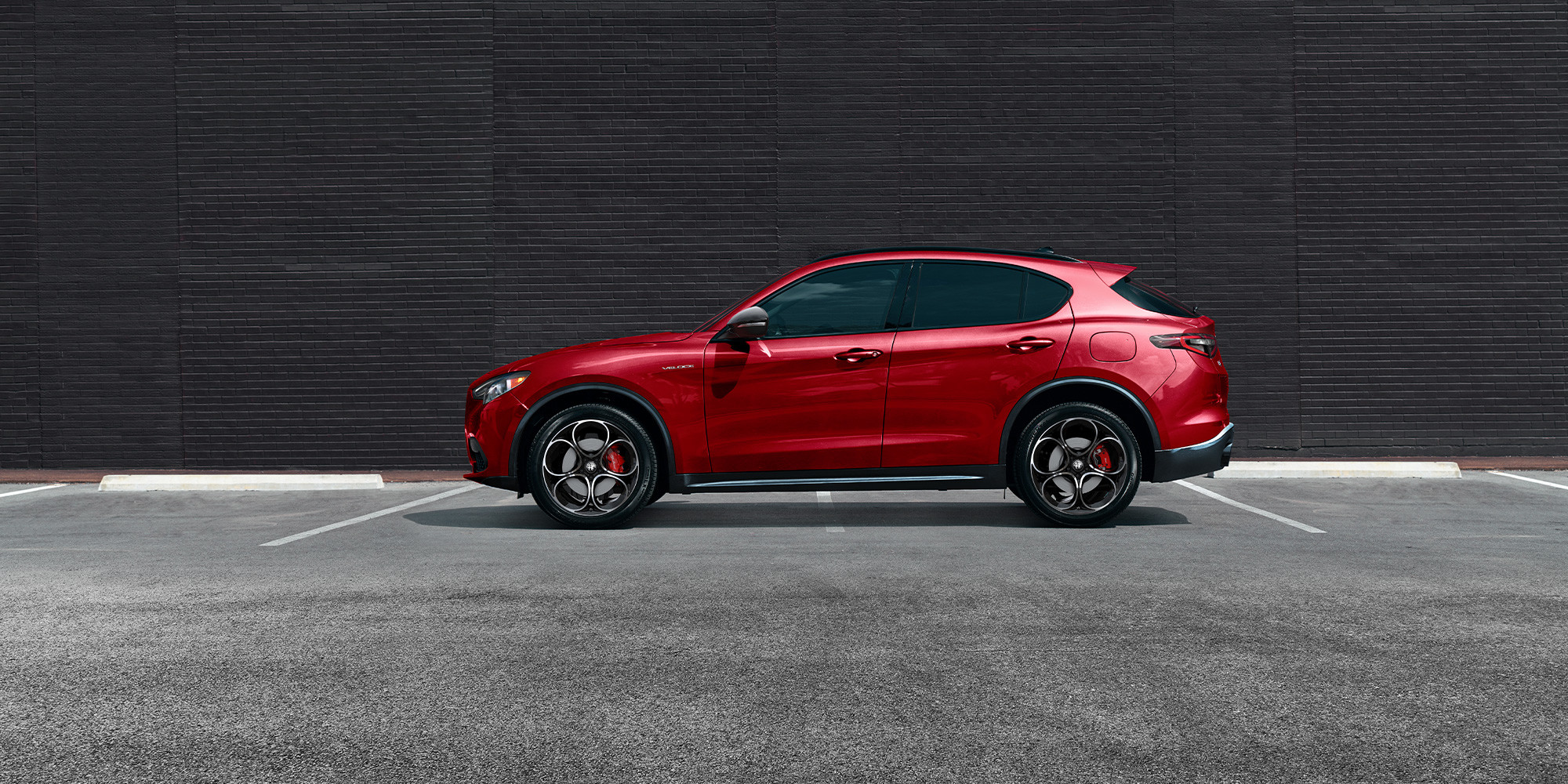 Vue latérale d'une Alfa Romeo Stelvio rouge 2025 garée contre un mur de briques sombres.