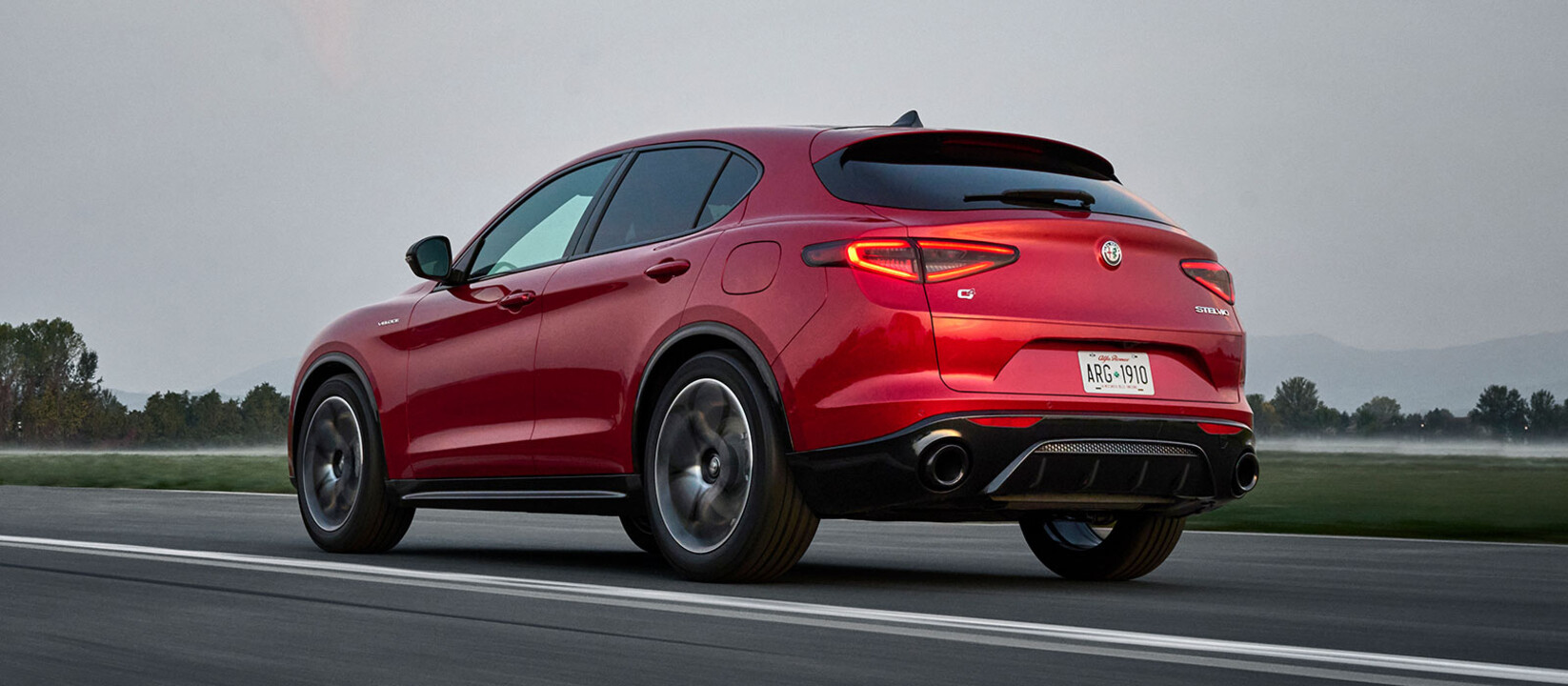 A back angled view of a red 2025 Alfa Romeo Stelvio seen driving on a paved roadway.