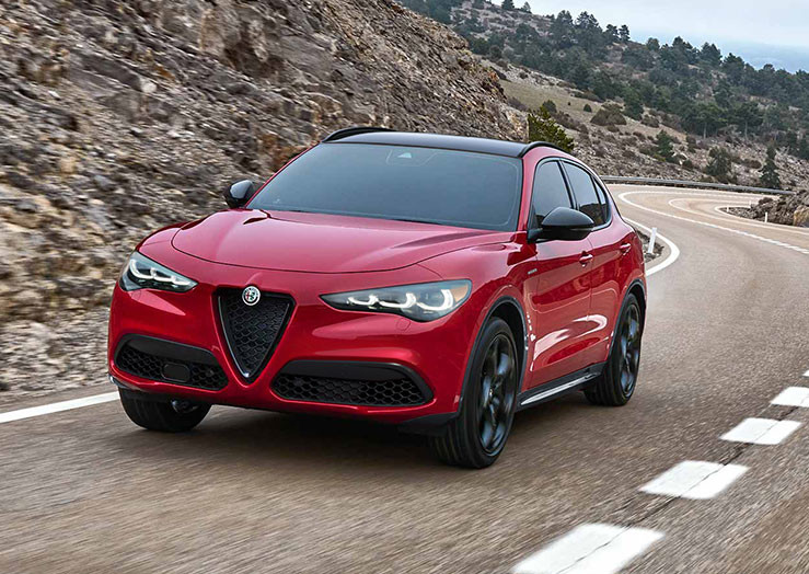 A front view of a red 2025 Alfa Romeo Stelvio shown driving on paved roadway with mountain terrain seen behind.
