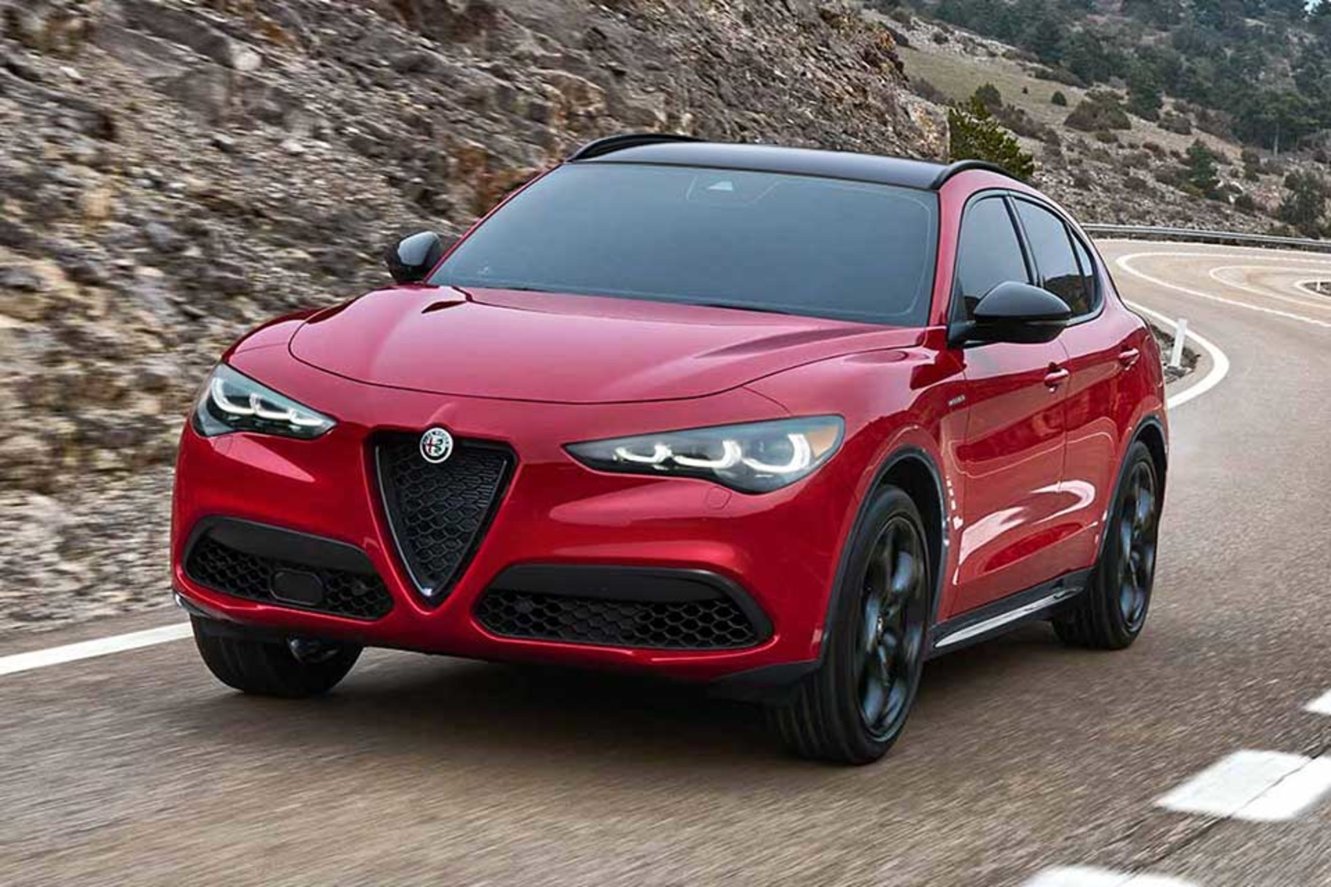 A red 2026 Alfa Romeo Stelvio is maneuvering around a mountain curve, showcasing its sporty design and agility on winding roads.