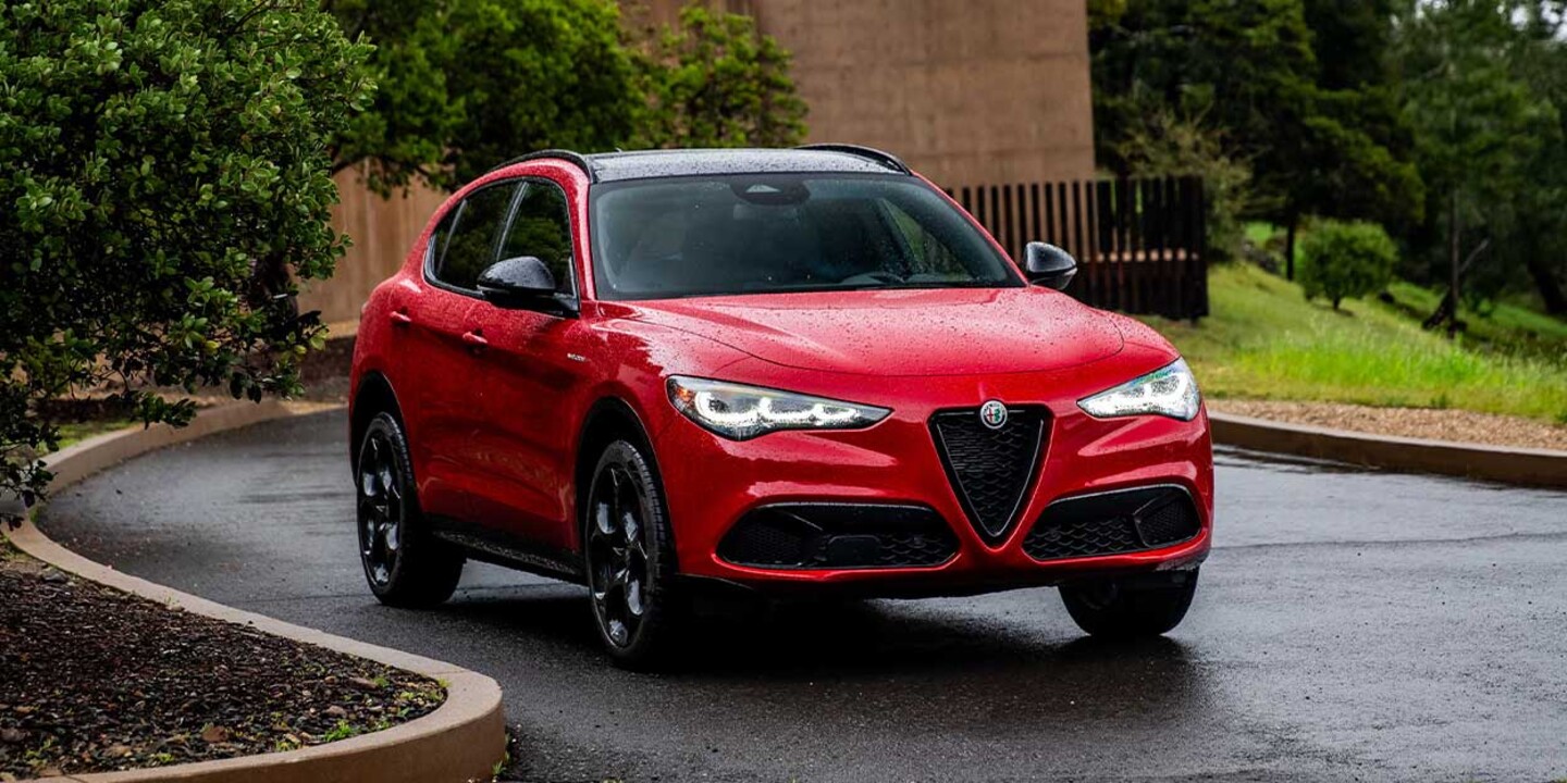 Red 2026 Alfa Romeo Stelvio parked on a wet driveway, with lush greenery and a stone wall in the background.