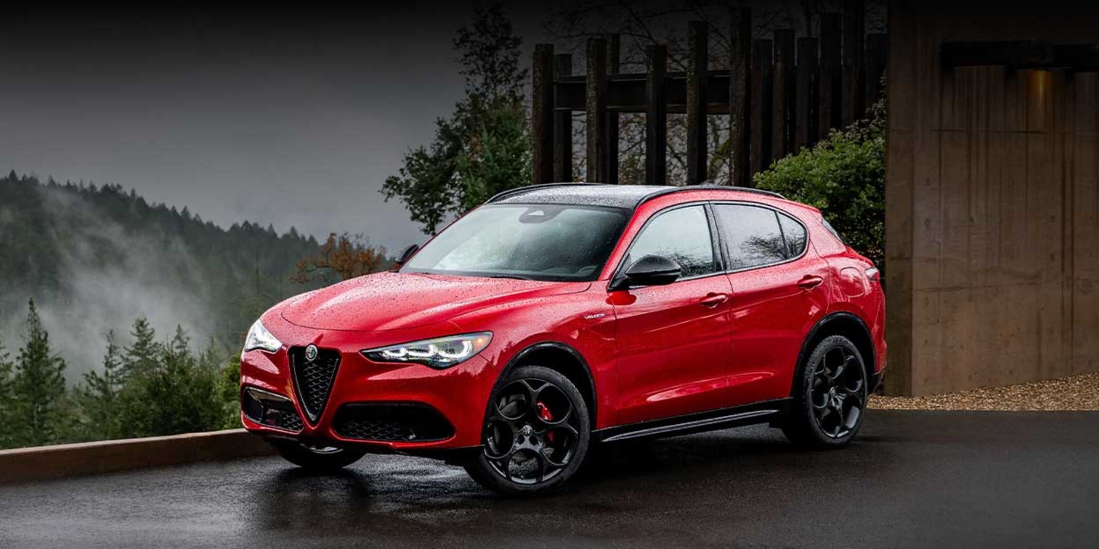 2026 Alfa Romeo Stelvio in red, parked on a wet road with lush green forests and misty mountains in the background.