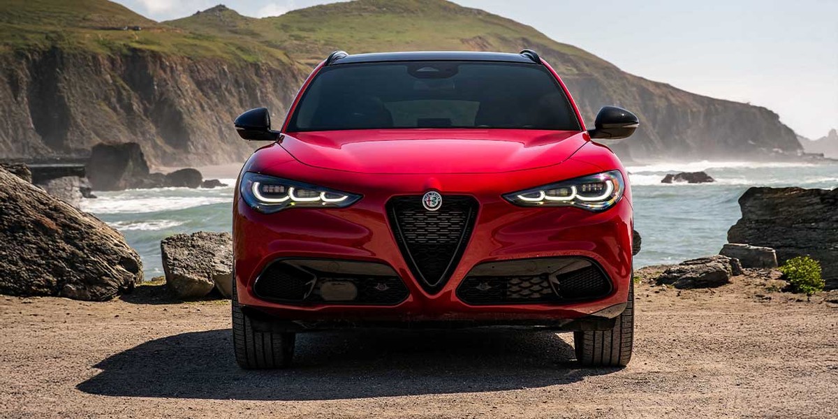 Front view of the 2026 Alfa Romeo Stelvio in red, parked on a coastal road with dramatic cliffs and ocean waves in the background.