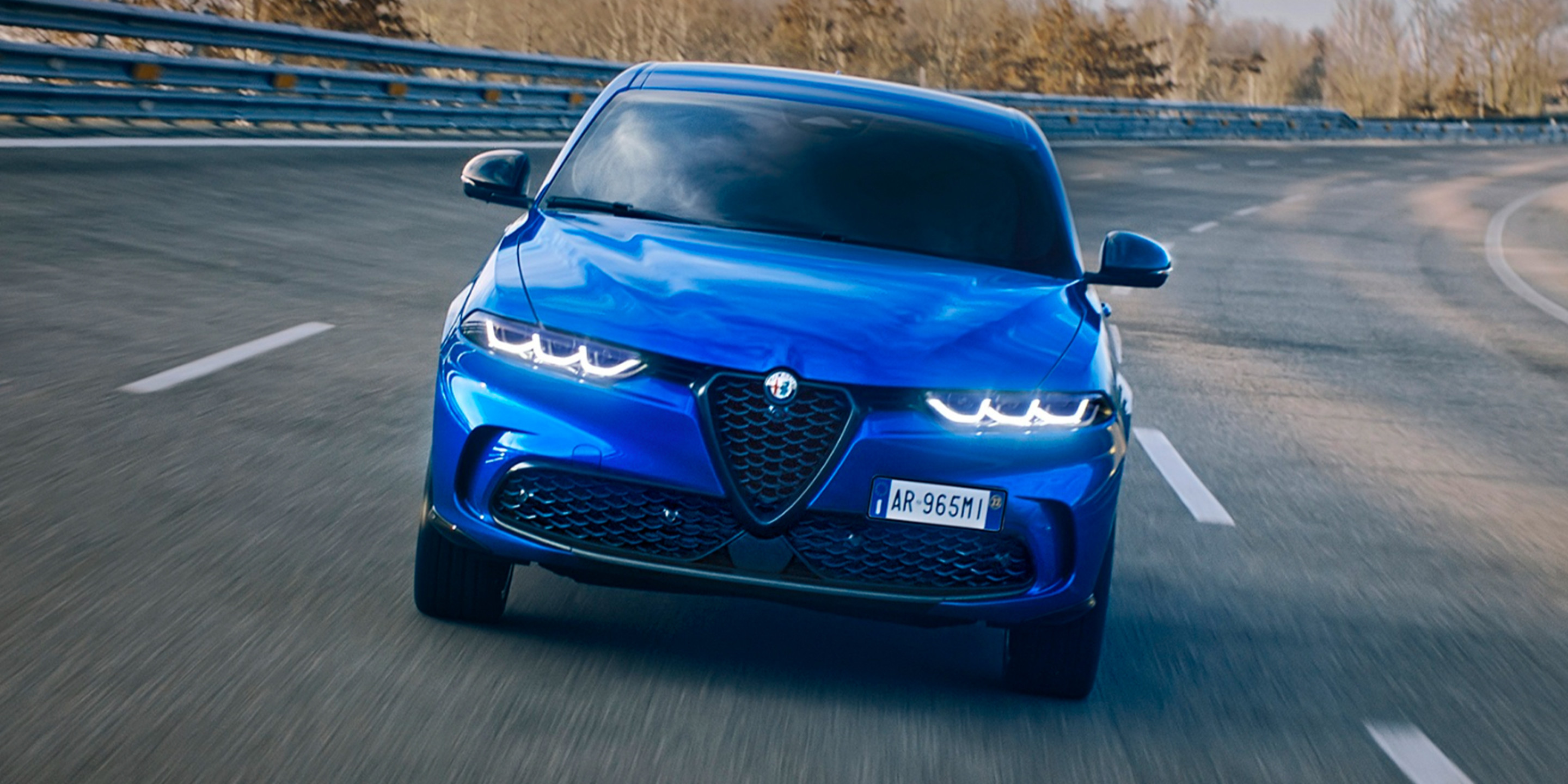 Alfa Romeo Tonale 2025 de couleur bleu Misano métallisé roulant sur une route incurvée avec des arbres et un ciel nuageux en arrière-plan.