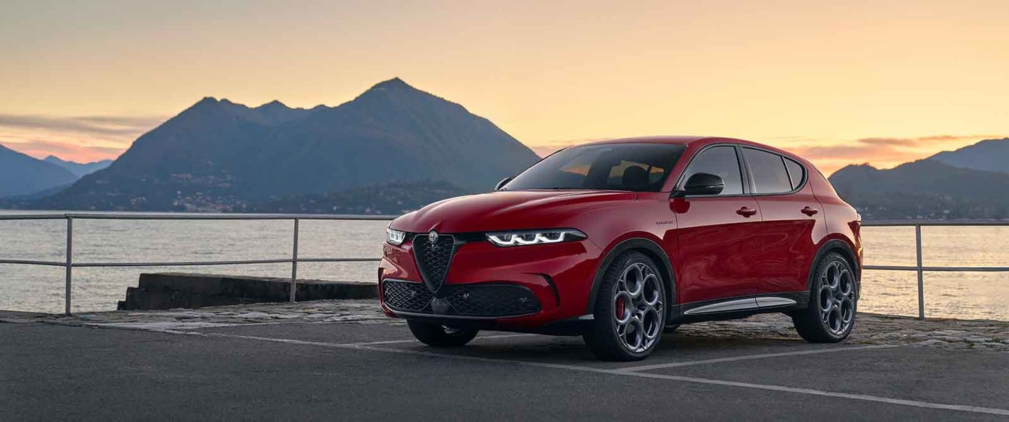 Alfa Romeo Tonale 2025 de couleur Alfa Rosso garée sur le trottoir avec vue sur un lac et des montagnes au loin éclairées par le lever du soleil.