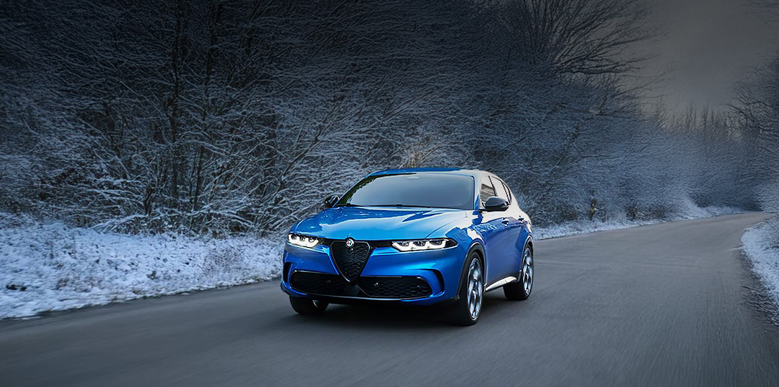 Une Alfa Romeo Tonale bleue 2025 roulant sur une route avec des arbres enneigés en arrière-plan.