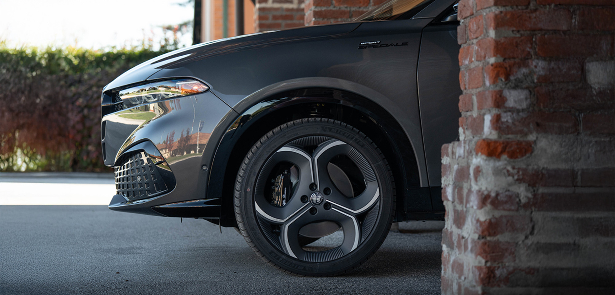 Vue latérale d'un Alfa Romeo Tonale à essence gris 2026 mettant en valeur le design élégant des jantes, se détachant sur un fond architectural en briques, soulignant son élégance et son style.