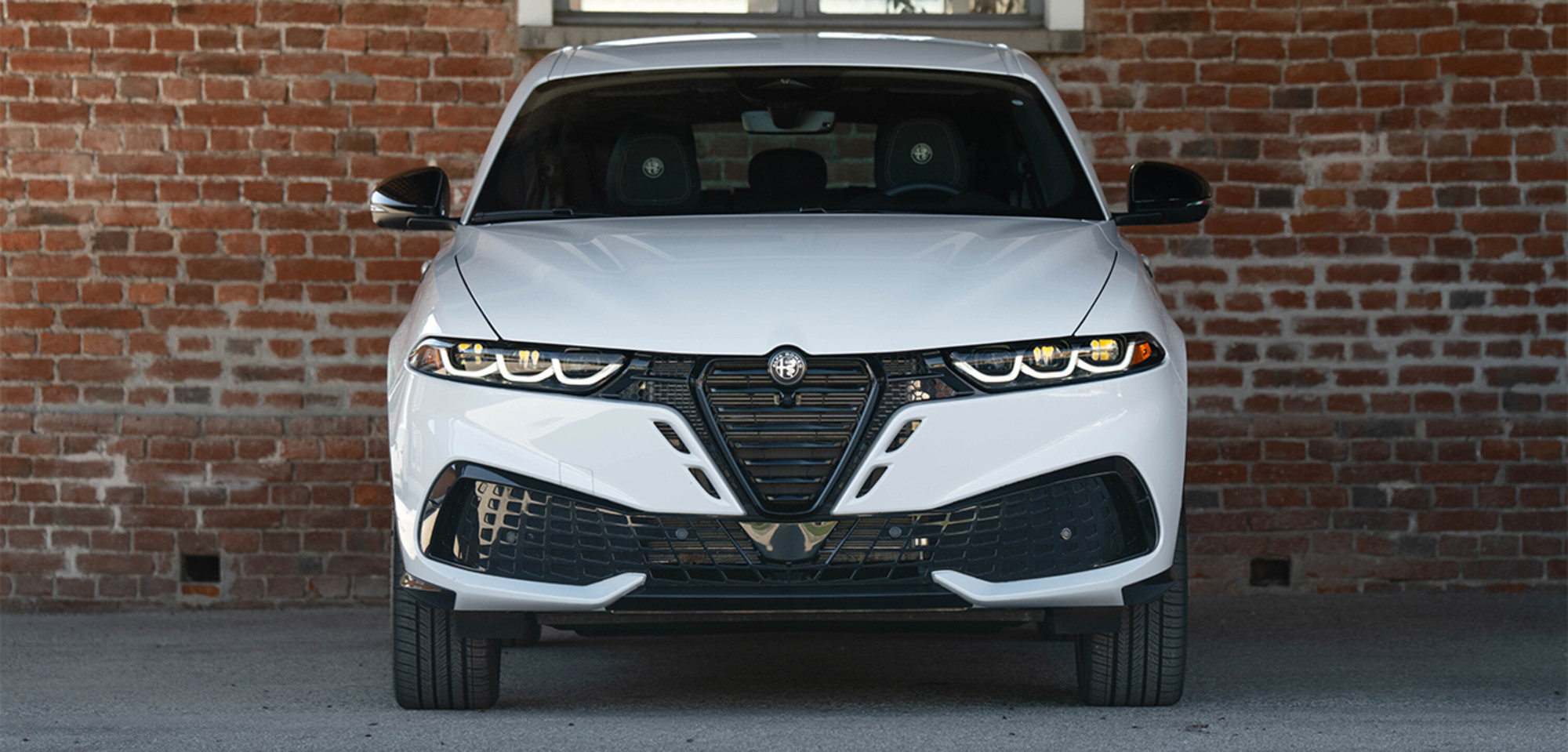 Vue de face d'un Alfa Romeo Tonale à essence blanc 2026, positionné contre un mur de briques, mettant en valeur sa calandre distinctive et ses phares élégants.