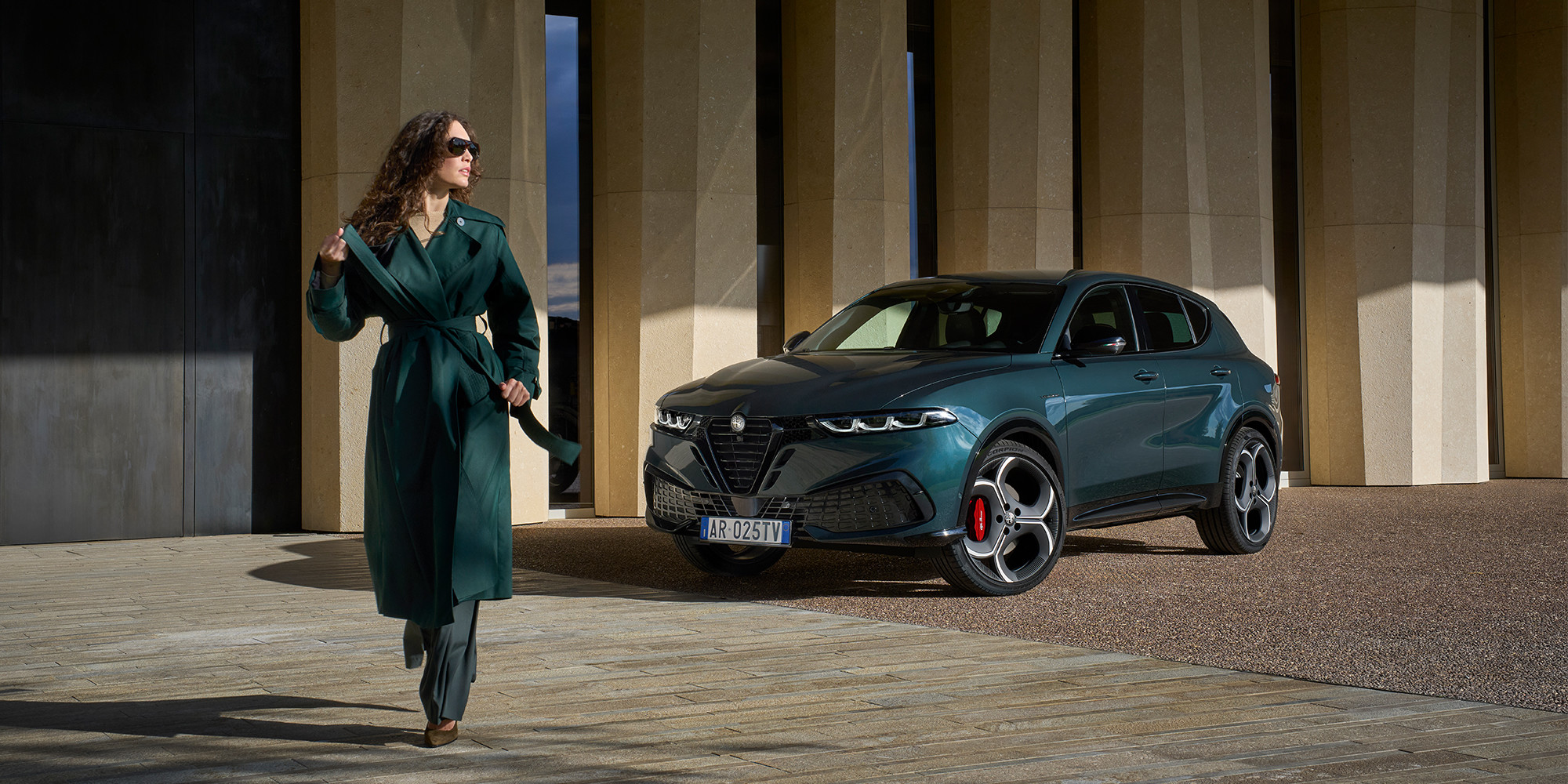 Un Alfa Romeo Tonale à essence 2026, couleur vert forêt, est élégamment garé devant un bâtiment d'architecture moderne, tandis qu'une personne vêtue d'une tenue assortie marche à proximité.