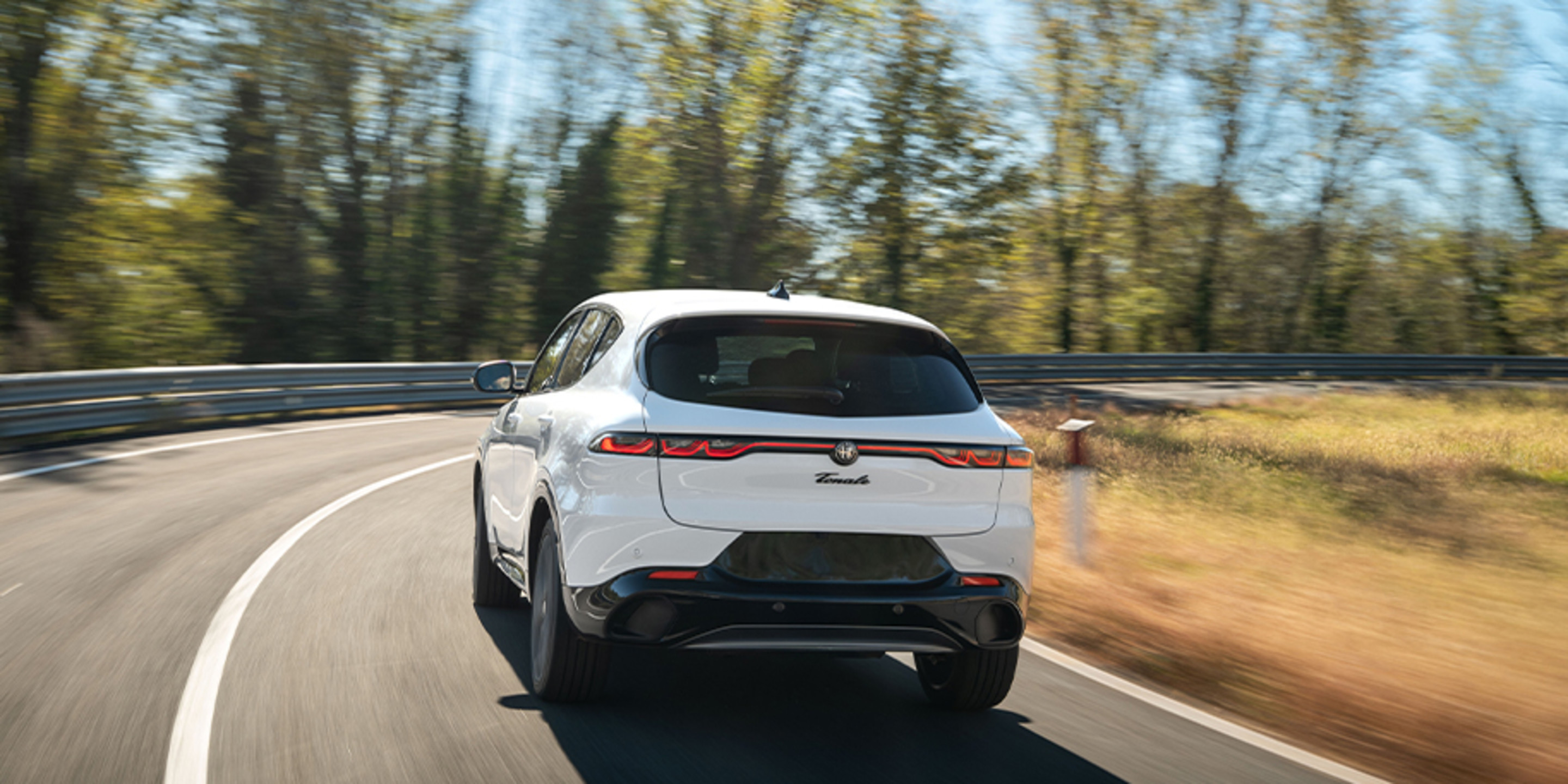 A white 2026 Alfa Romeo Tonale Gas driving on a winding forest road, highlighting its sleek rear design and dynamic performance.