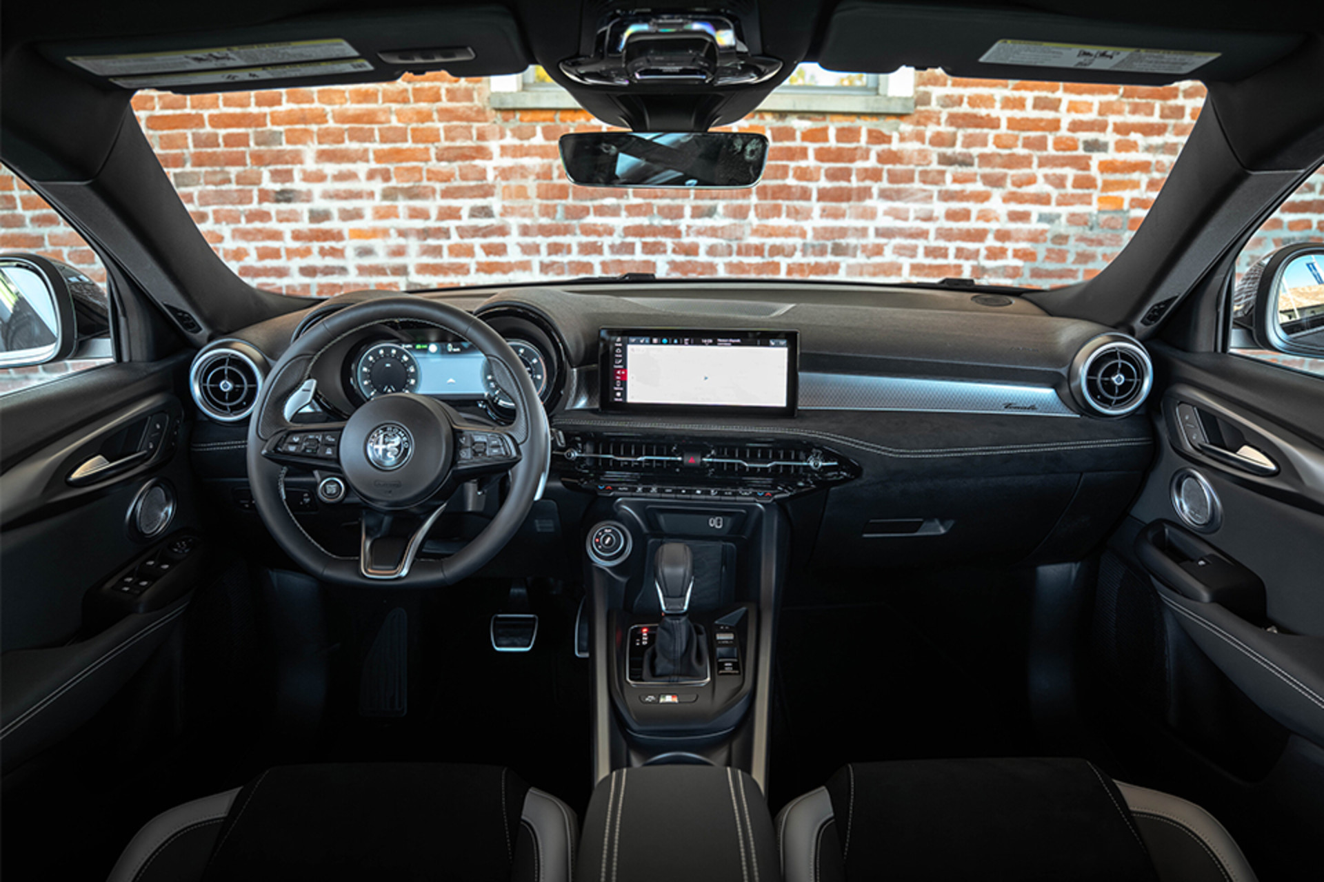 Intérieur d'un Alfa Romeo Tonale à essence 2026 présentant un tableau de bord moderne avec un volant élégant, des affichages numériques et une disposition sophistiquée des commandes sur fond de mur de briques.