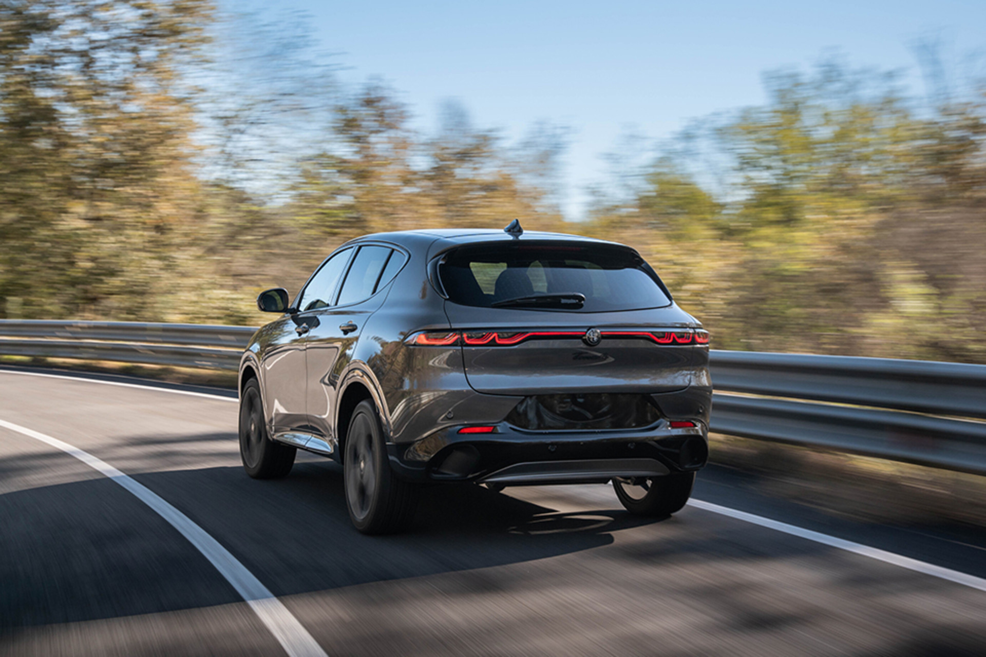 Un Alfa Romeo Tonale à essence gris 2026 circulant sur une route sinueuse, mettant en valeur son design arrière élégant et son profil sportif dans un décor boisé.