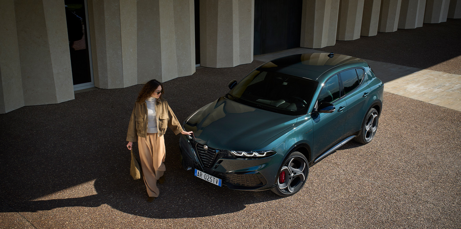 Une femme marche à côté d'un Alfa Romeo Tonale à essence 2026 de couleur vert forêt, garé devant un bâtiment d'architecture moderne.