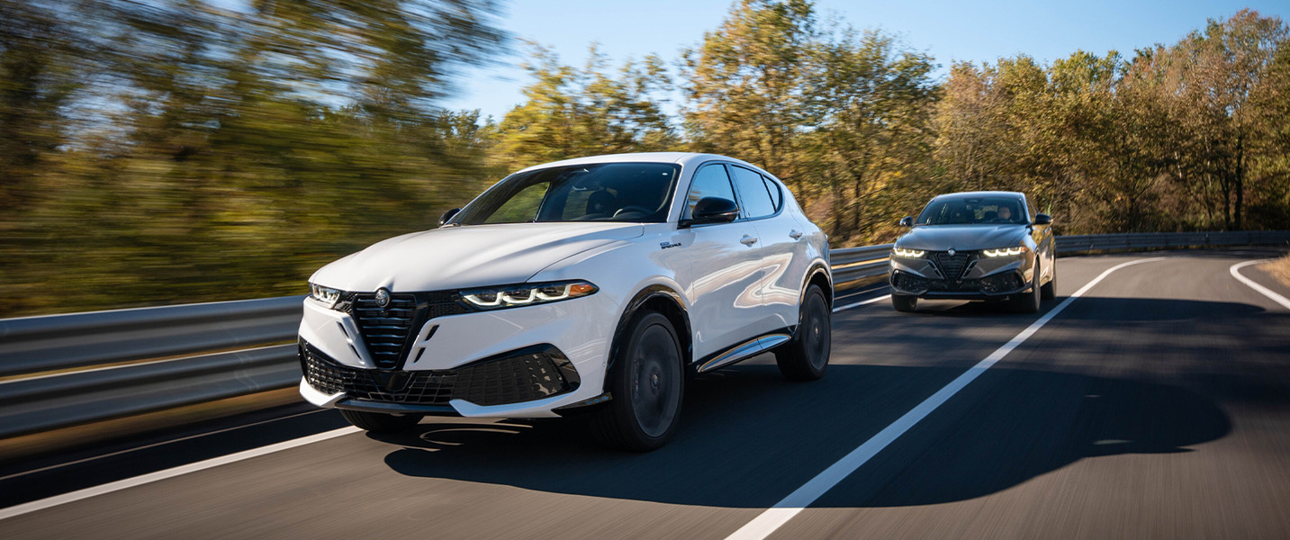 A white 2026 Alfa Romeo Tonale Gas driving on a forested road, with another vehicle following, highlighting its sleek design and dynamic performance.
