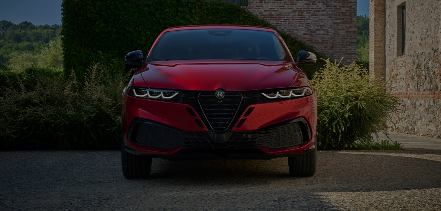 Un Alfa Romeo Tonale à essence rouge 2026 garé sur une allée en pierre, encadrée par de la verdure et un bâtiment en briques, mettant en valeur son design avant audacieux.