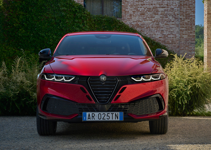 Alfa Romeo Tonale essence 2026 rouge stationné sur une allée en pierre bordée de verdure, près d’un bâtiment en brique, mettant en valeur son design avant audacieux.