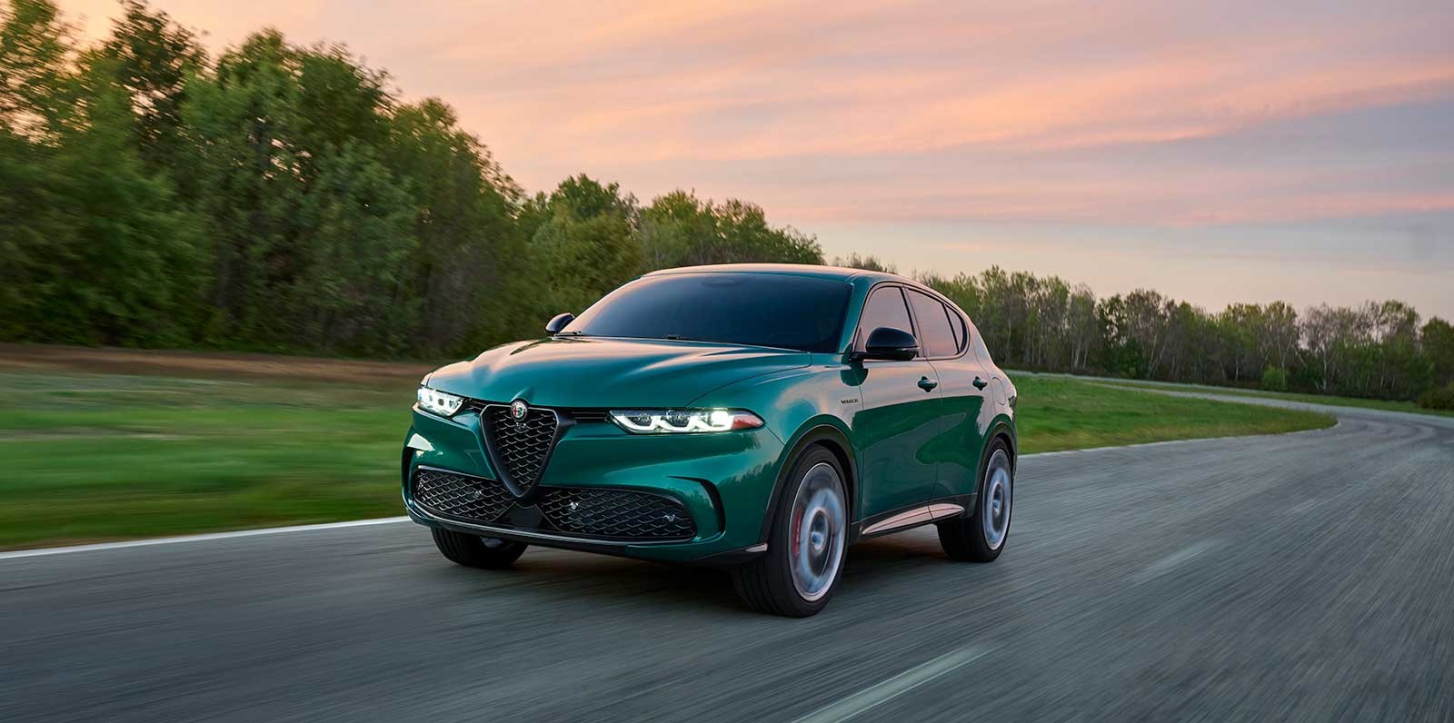 Alfa Romeo Tonale Veloce VHR vert 2025 roulant sur une route au coucher du soleil avec des arbres et de la verdure en arrière-plan.