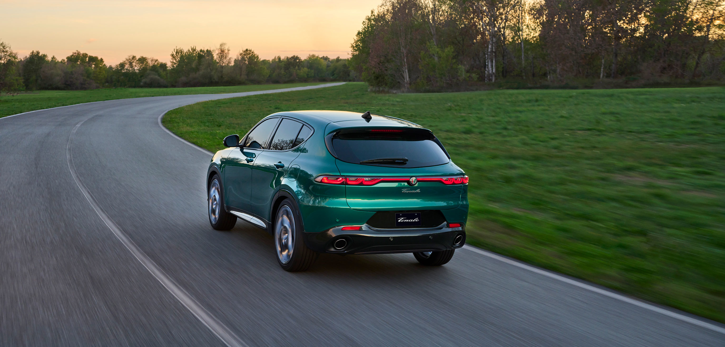 Vue arrière d'un Alfa Romeo Tonale VHR Veloce 2025 roulant sur une route sinueuse entourée de verdure au coucher du soleil.