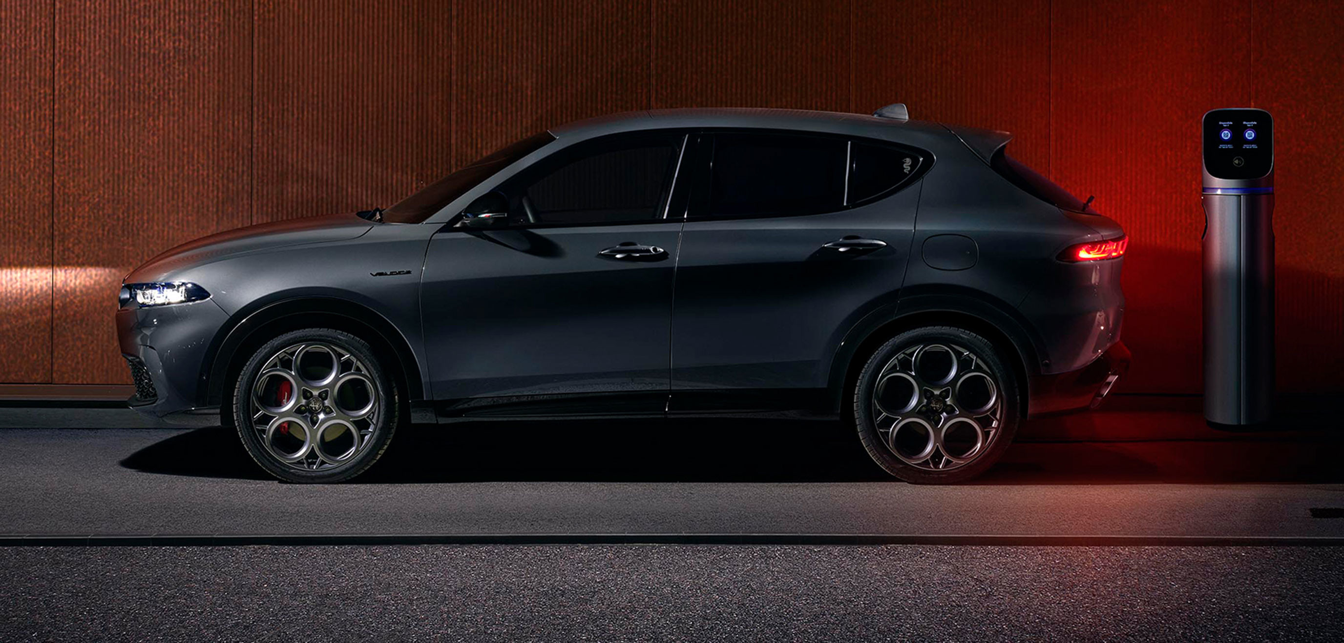 Vue latérale d'un Alfa Romeo Tonale VHR de couleur Grigio Ascari métallisé 2025 garé à côté d'une borne de recharge.