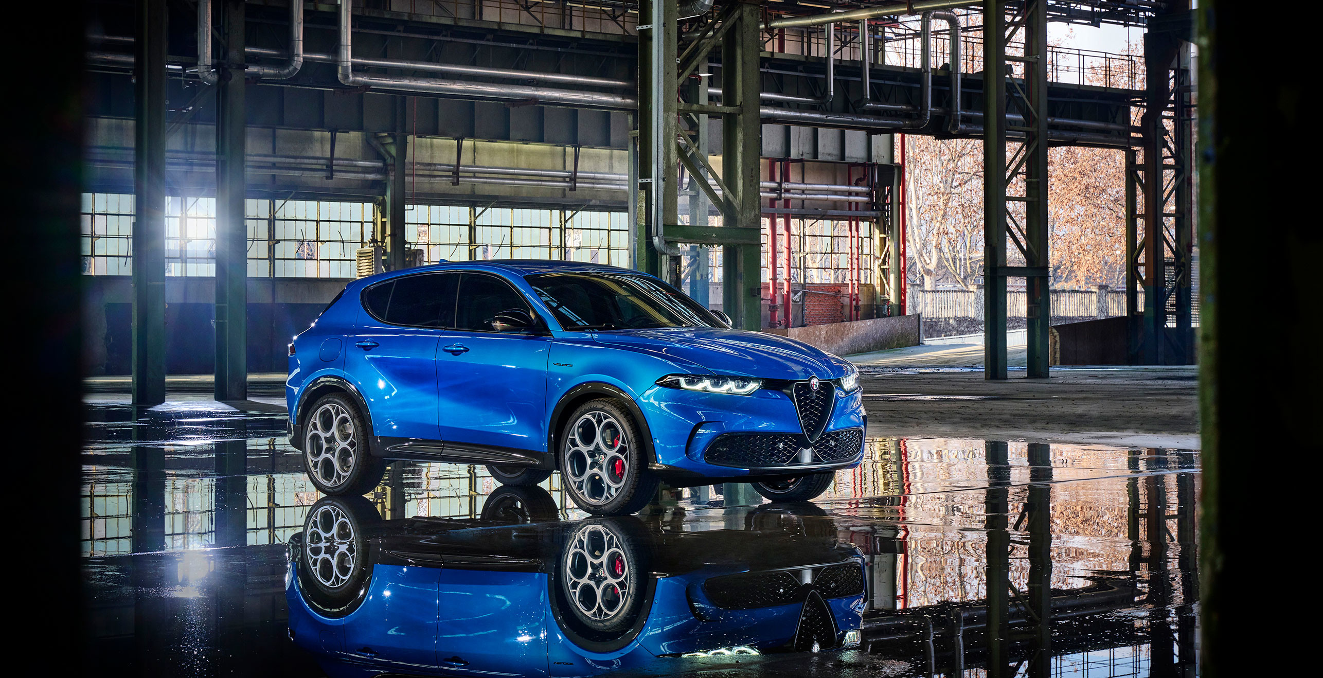 Alfa Romeo Tonale PHEV 2025 de couleur bleu Misano métallisé garé à l'intérieur d'un bâtiment industriel avec des reflets sur le sol mouillé.