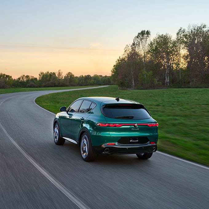 Rear view of a 2025 Alfa Romeo Tonale PHEV Veloce driving on a winding road surrounded by greenery at sunset.