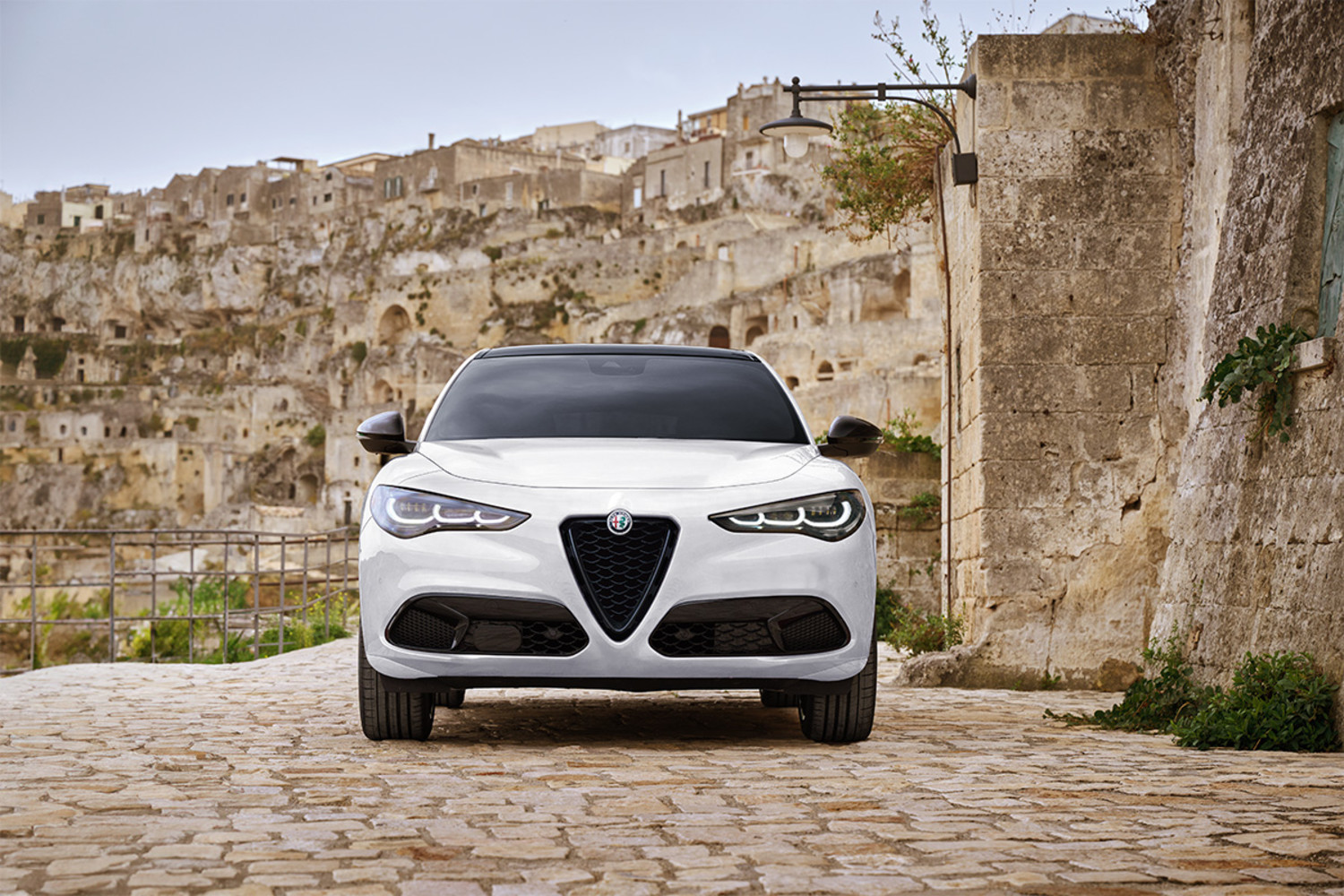 Vue avant d’un Alfa Romeo Stelvio Tributo Italiano 2025 en blanc Alfa, stationné dans une rue pavée avec des bâtiments anciens en pierre en arrière-plan.