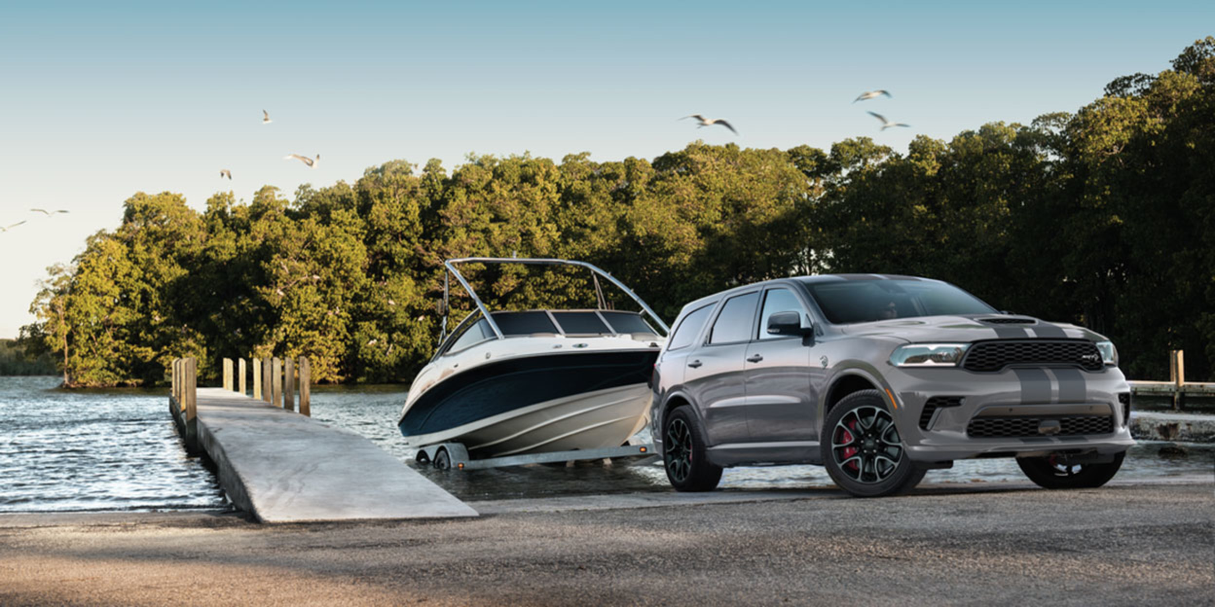 The side profile view of a grey 2023 Dodge Durango towing a white boat out of a body of water next to a dock with trees shown behind.