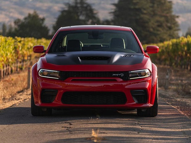 The front view of a red 2023 Dodge Charger parked on paved road in between rows of green plants and trees shown in the distance.