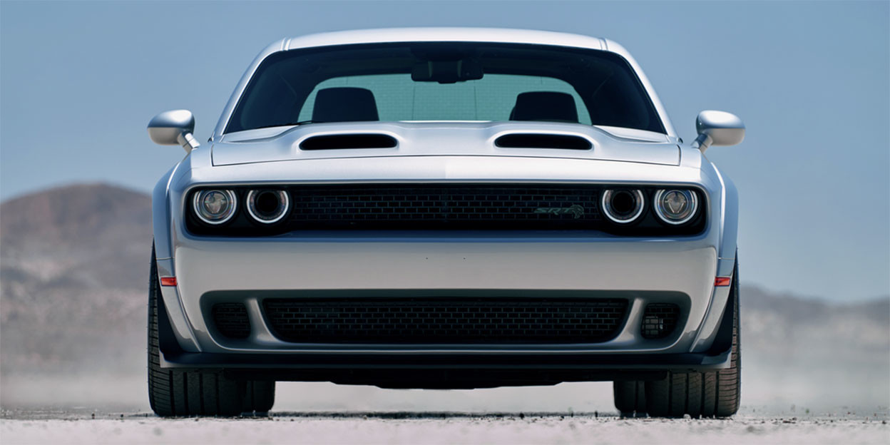 The front view of a silver 2023 Dodge Challenger parked on unpaved ground with mountains and a blue sky shown behind. Schedule a Sales Appointment