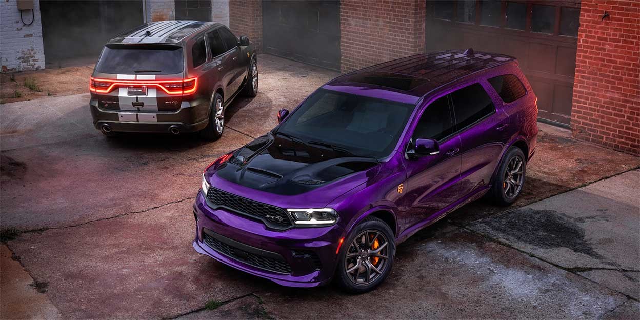 Deux véhicules Dodge Durango 2026 garés dans une zone industrielle, l'un violet avec une vue de face et l'autre gris avec une vue de dos, devant des bâtiments en briques. 