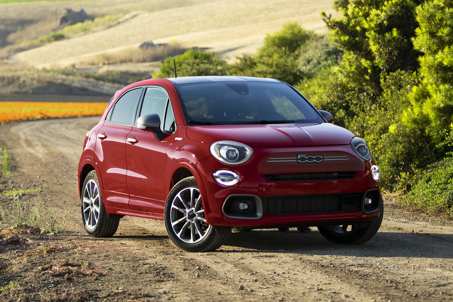 Front view of a red 2023 Fiat 500X parked on a dirt road with vast land and trees in the background.