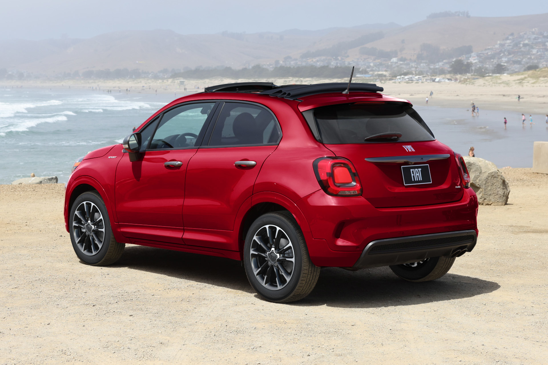 Profile view of a red 2023 Fiat 500X parked on a sandy path with a beach shown in the background.