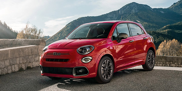Side view of a red 2023 Fiat 500X parked near a stone wall with mountains in the background.